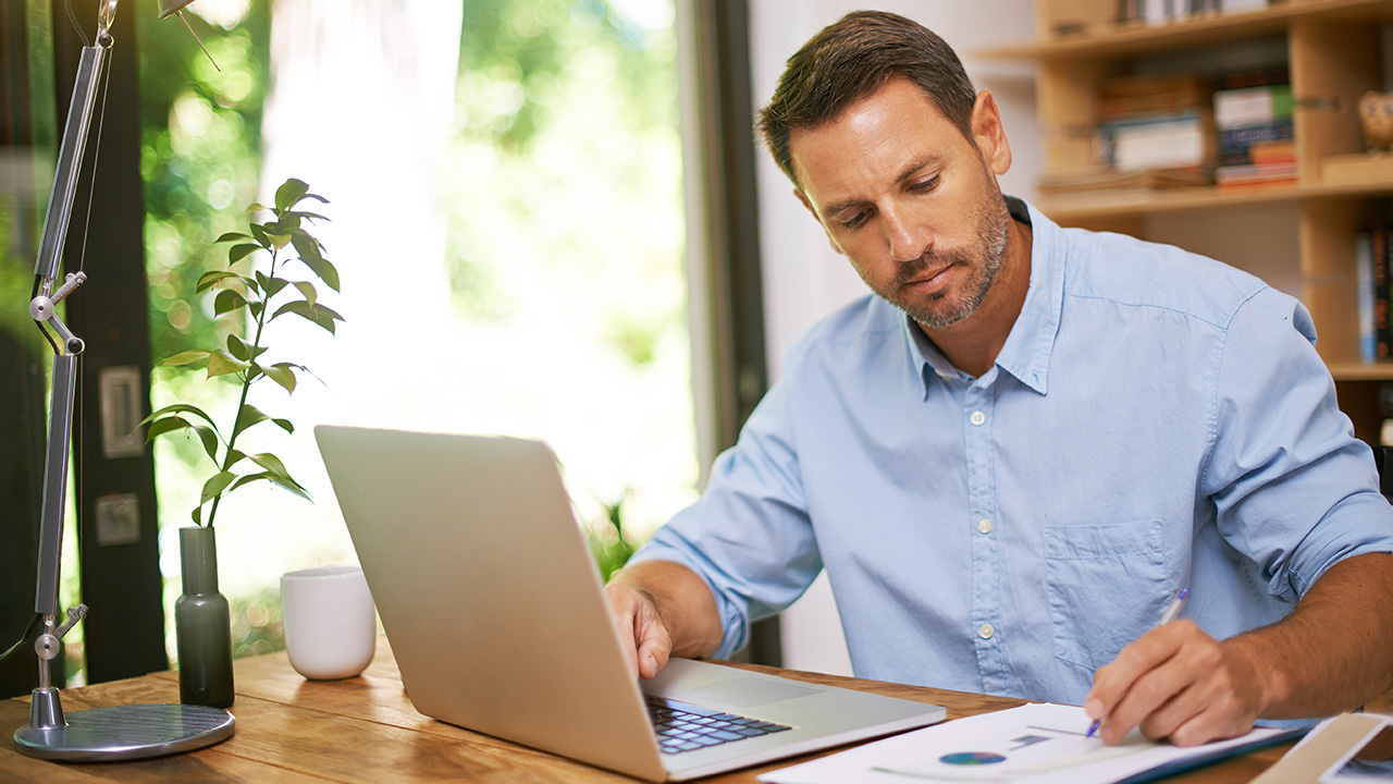 A man writing down on his paper while working on his laptop